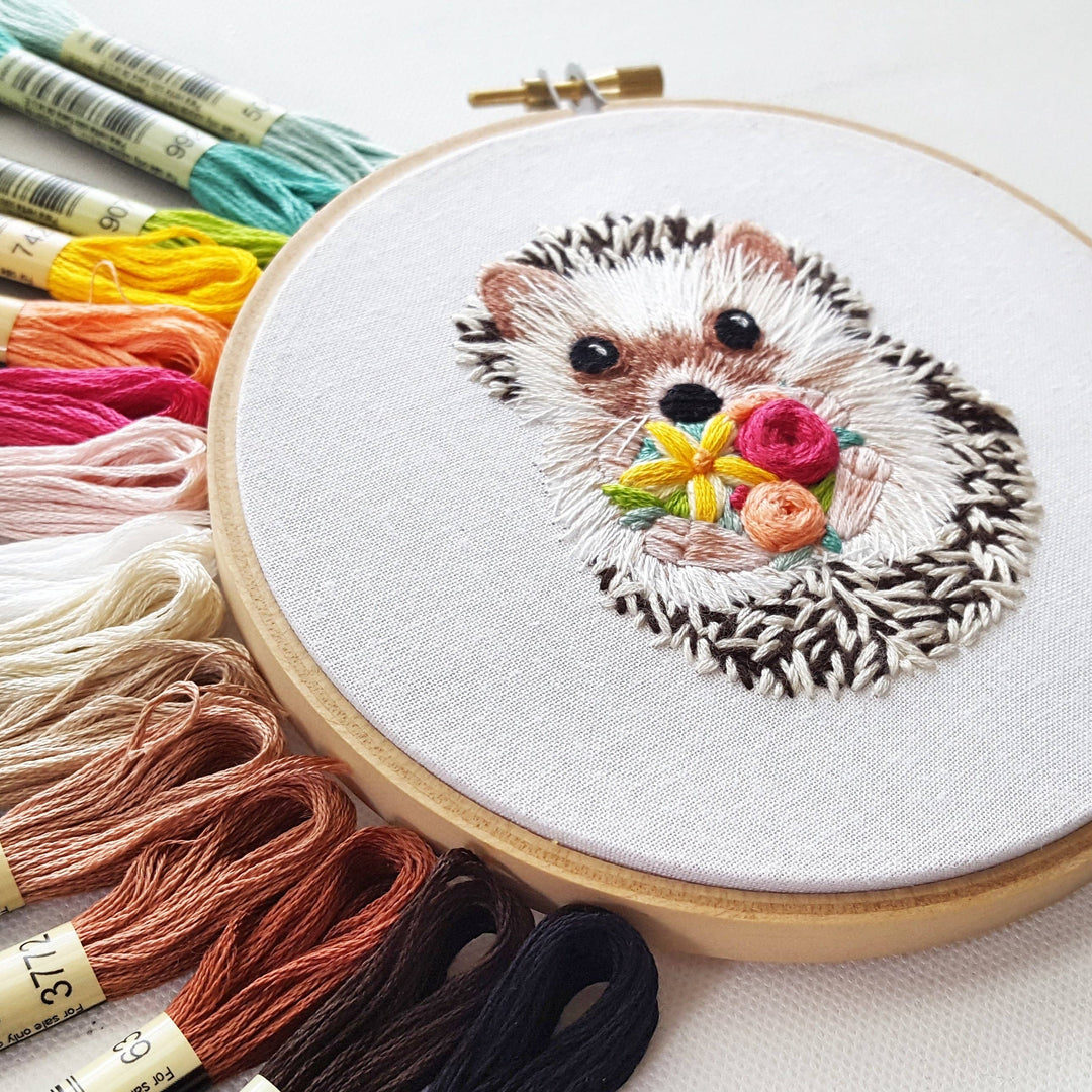 Embroidery hoop with a hedgehog holding flowers surrounded by colorful embroidery threads.