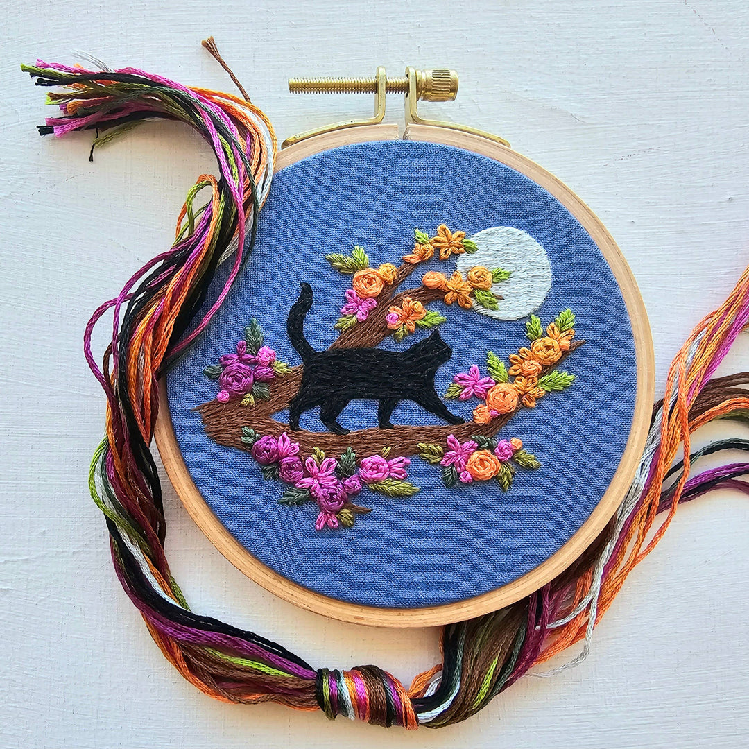 Embroidery hoop with a black cat and floral design on blue fabric, surrounded by colorful threads.