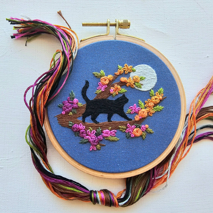 Embroidery hoop with a black cat and floral design on blue fabric, surrounded by colorful threads.