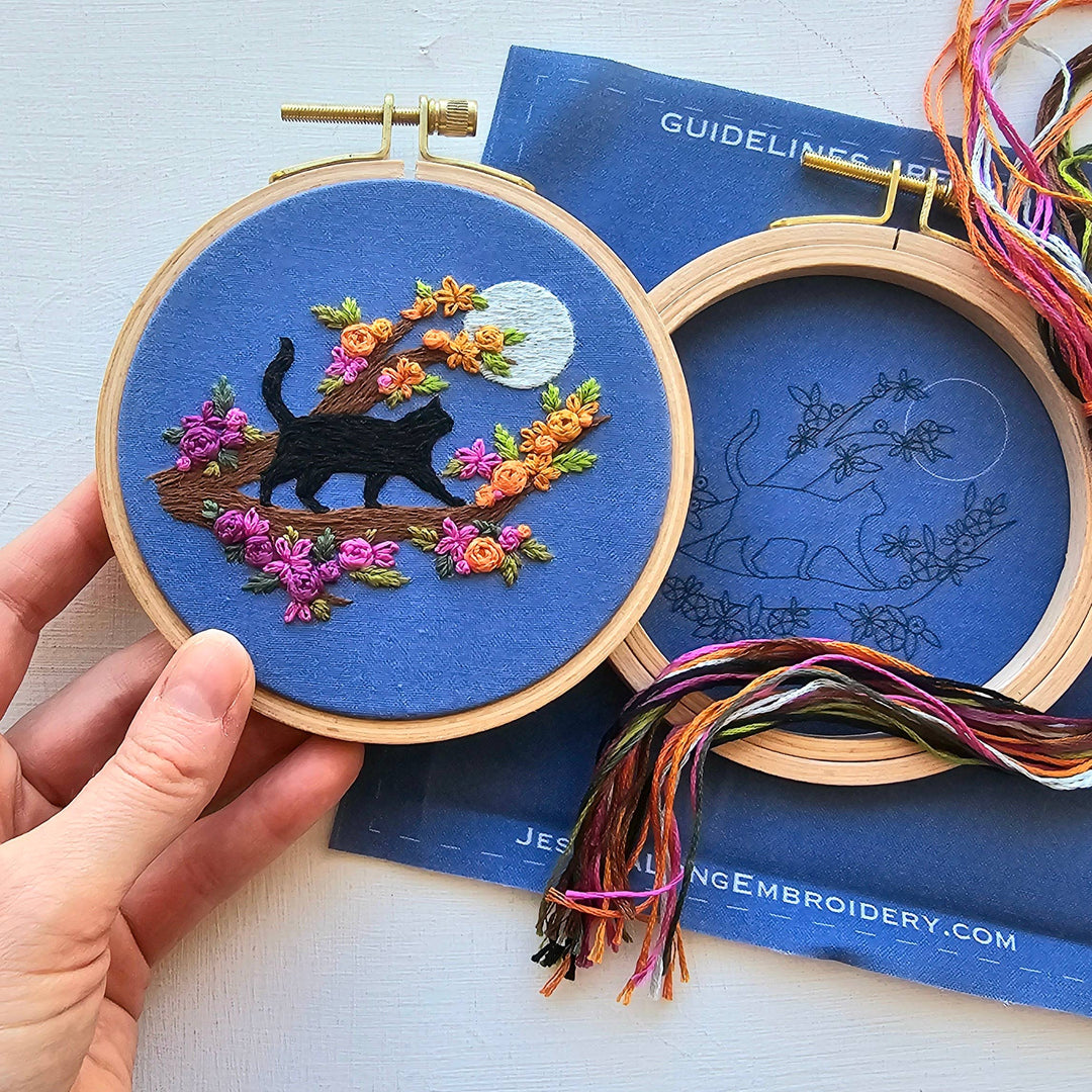 Embroidery hoop with a black cat design on a blue background, held by a hand.
