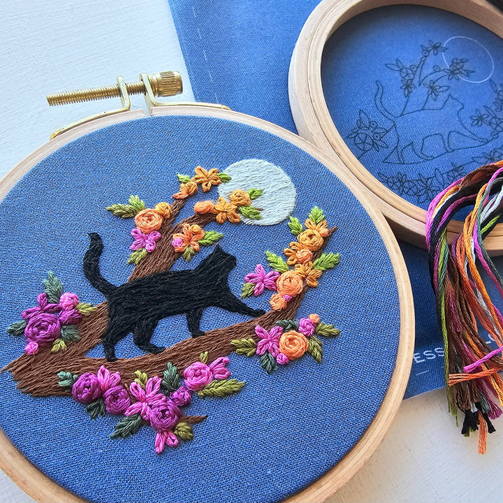 Embroidery hoop with a black cat and floral design on blue fabric, with another hoop and thread in the background.