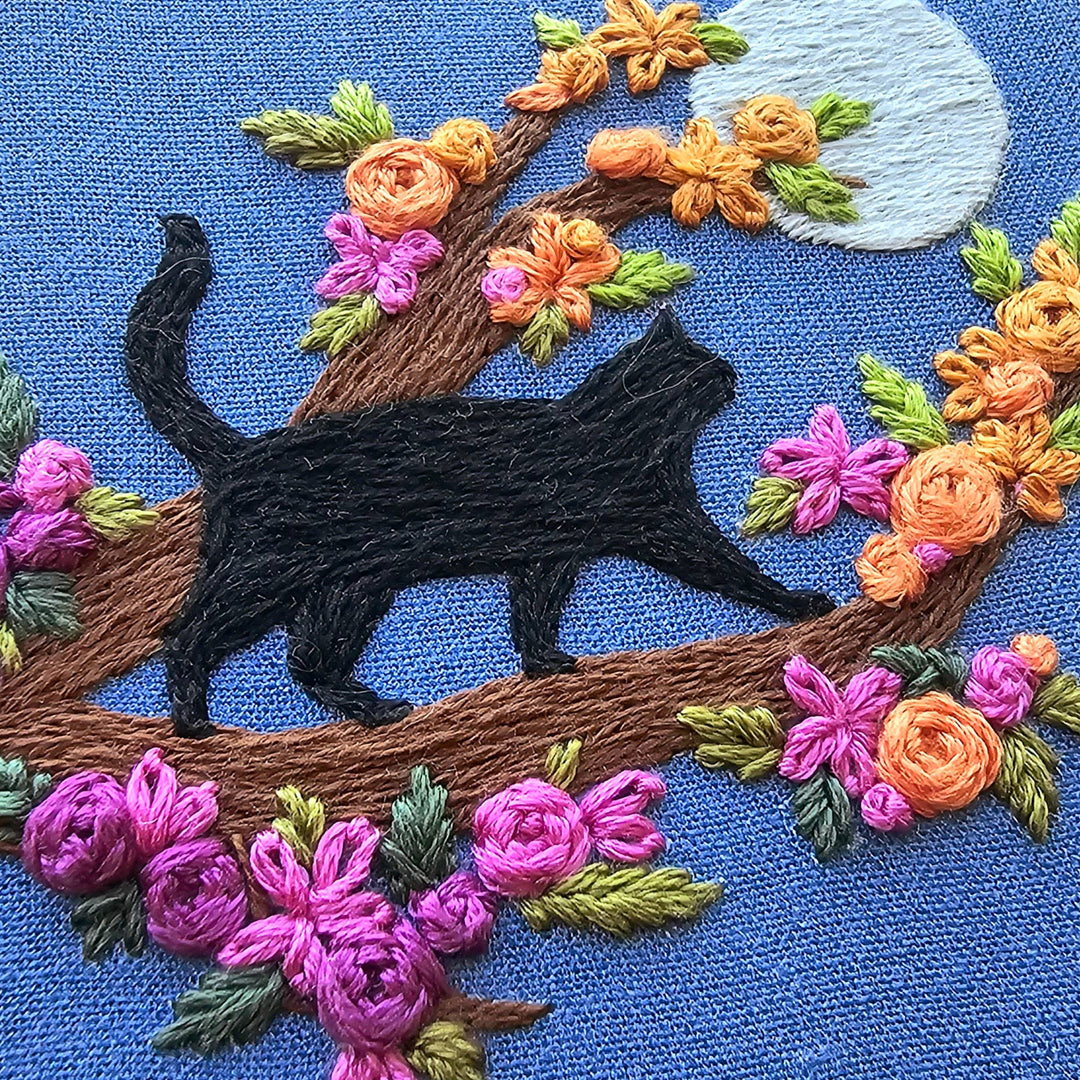 Embroidered design of a black cat on a branch with flowers on a blue background