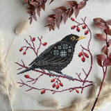 Cross-stitched bird on a branch with berries on a white background