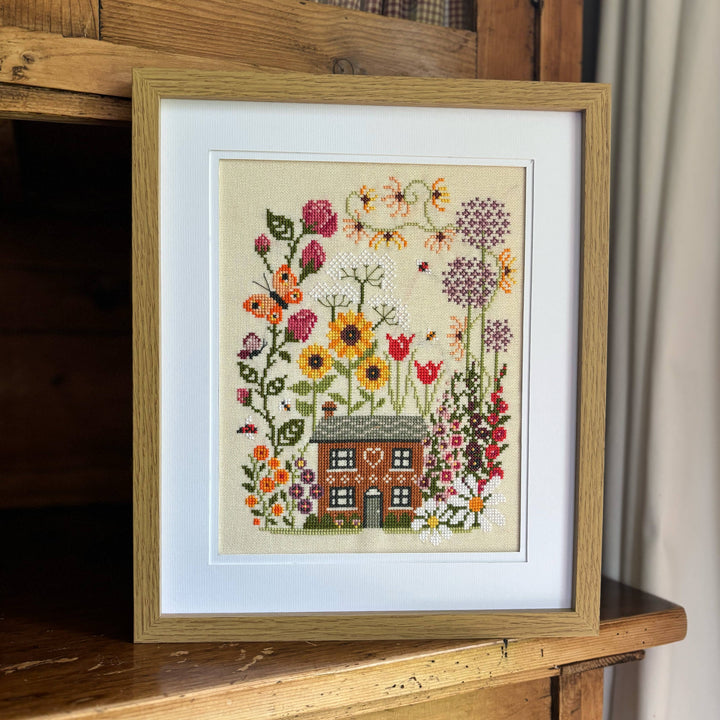 Framed cross-stitched artwork of a house surrounded by flowers on a wooden surface.
