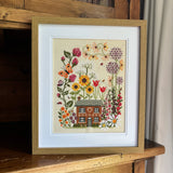 Framed cross-stitched artwork of a house surrounded by flowers on a wooden surface.