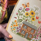 Cross-stitched design of a house with flowers and a butterfly, held by a person.