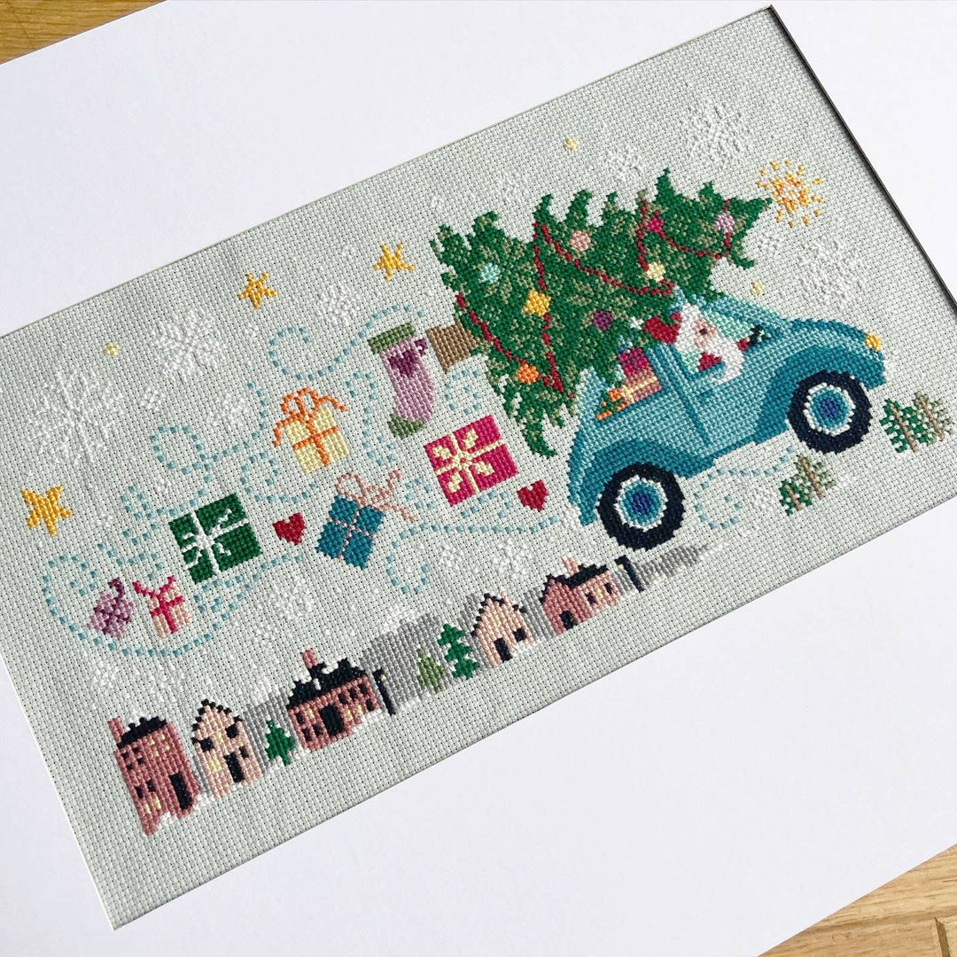 Cross-stitched panel with a blue truck carrying a Christmas tree and festive elements on a light background.