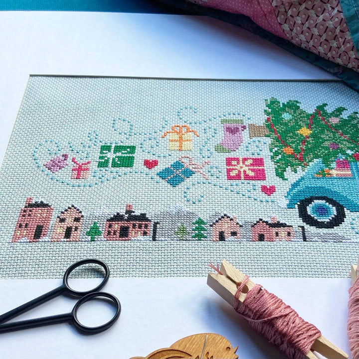 Cross-stitched scene with a truck, houses, and trees on fabric with scissors and thread nearby.