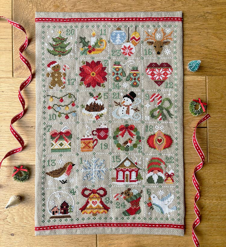 Cross-stitched Christmas-themed sampler on a wooden surface with decorative ribbons and small ornaments.