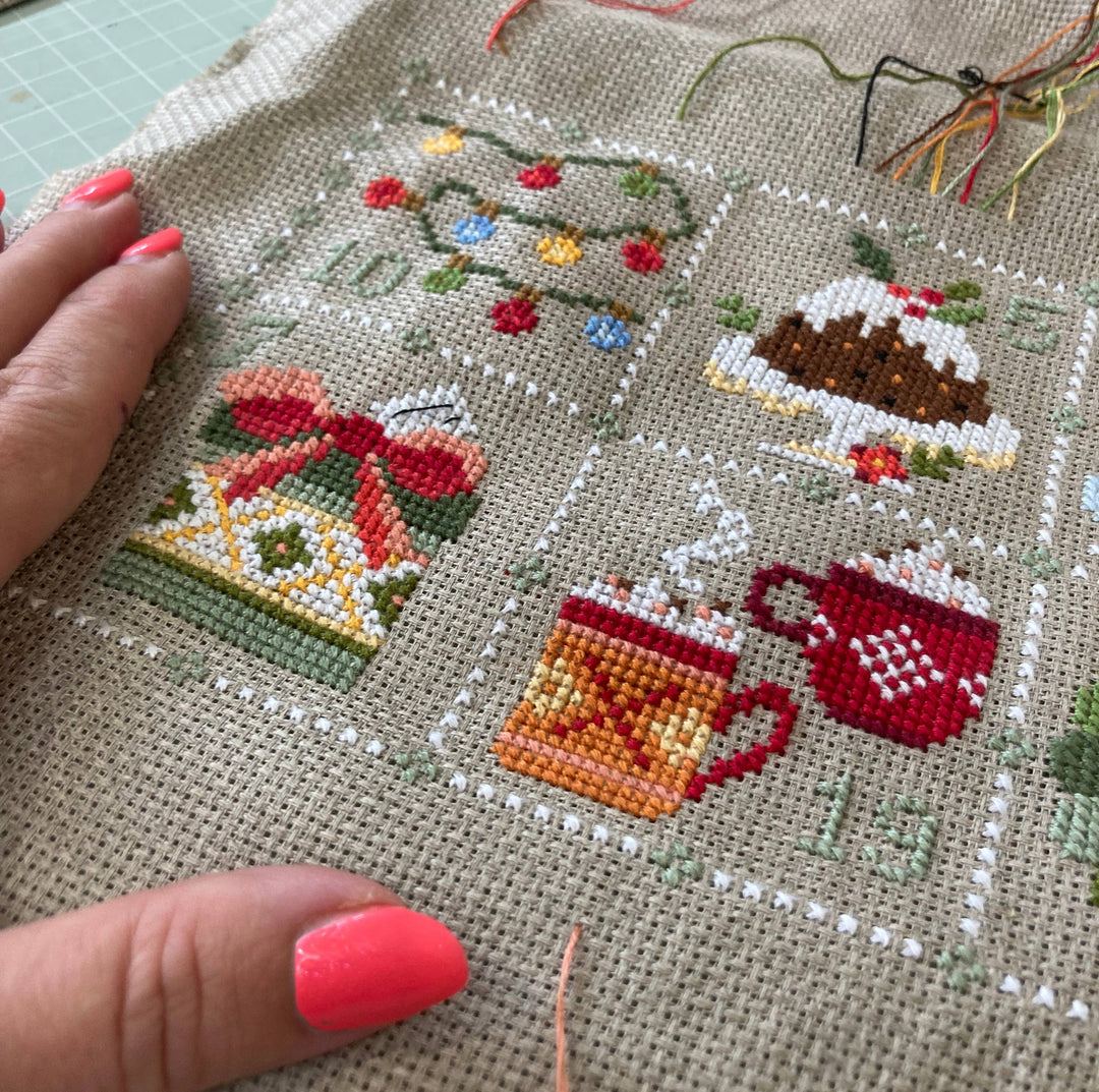 Cross-stitched piece with festive designs including a cake, mugs, and a bow.