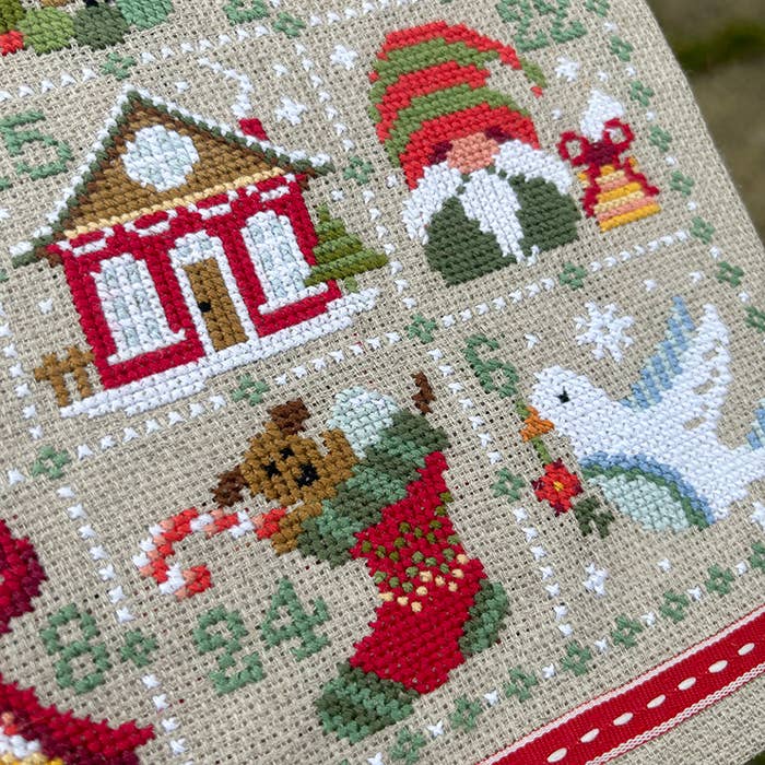 Cross-stitched Christmas scene with a house, gnome, dog, and stocking on a beige background.