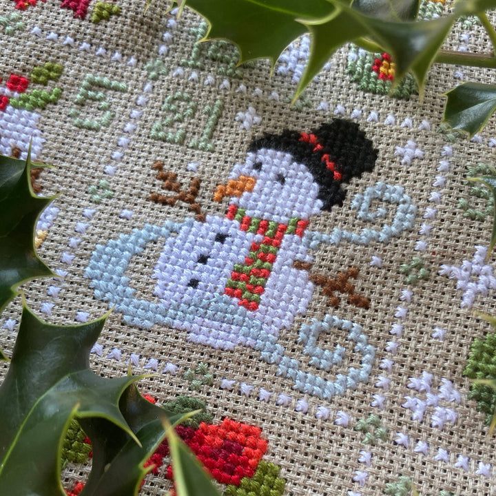 Cross-stitched snowman design on fabric with holly leaves around