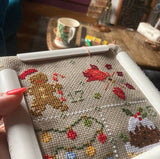 Cross-stitched gingerbread man and festive elements on fabric with a cup and saucer in the background.
