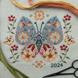 Cross-stitched butterfly design with floral patterns, surrounded by embroidery threads and scissors on a light background.