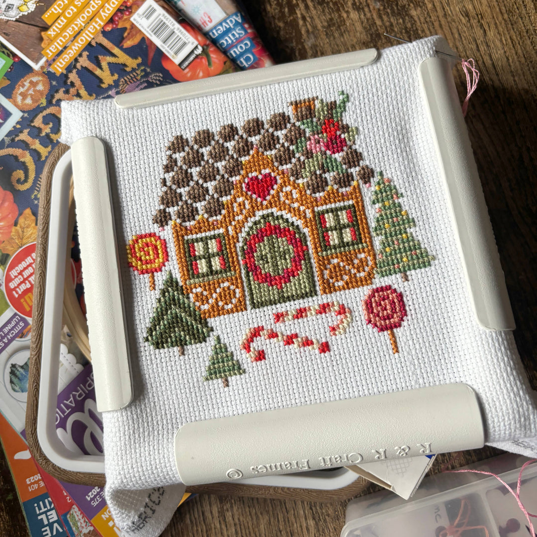 Cross-stitched gingerbread house on aida fabric with tools on a wooden surface