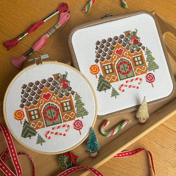 Cross-stitched gingerbread house design on fabric and hoop with embroidery threads on a wooden surface.