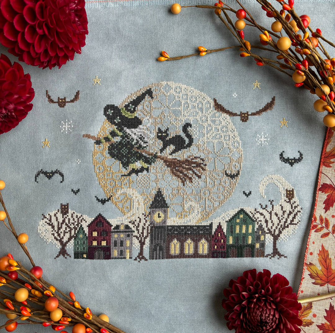 Cross-stitched design of a witch flying on a broomstick over a village with autumn decorations.