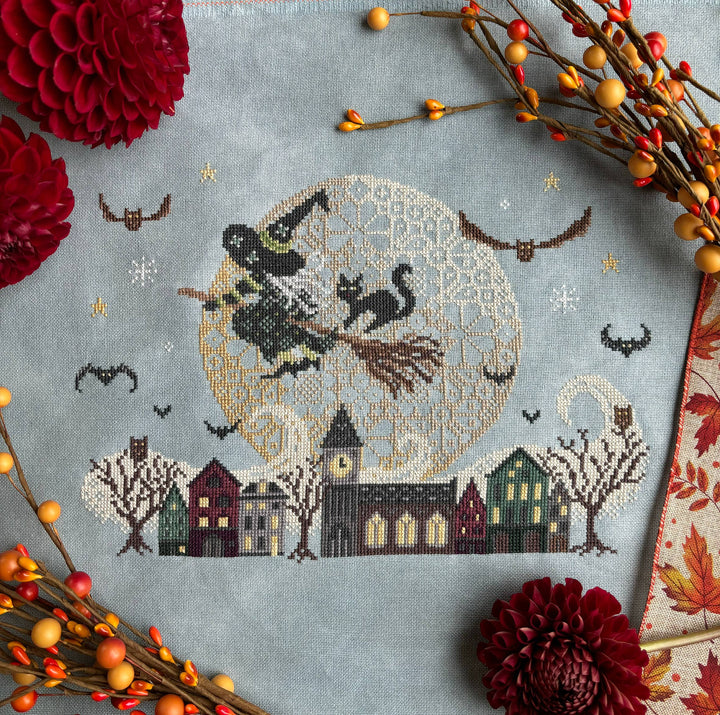 Cross-stitched design of a witch flying on a broomstick over a village with autumn decorations.