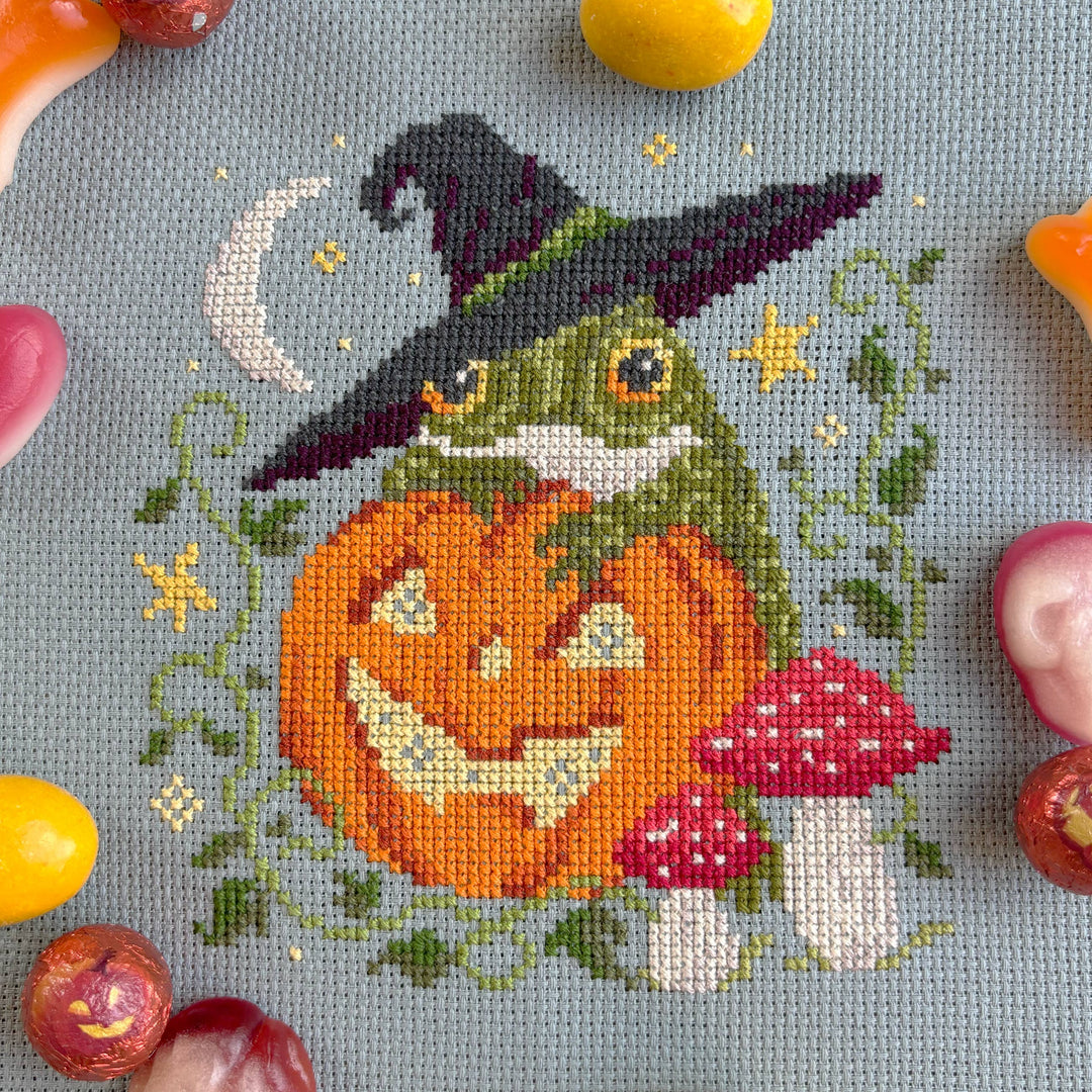 Cross-stitched design of a frog with a witch hat, pumpkin, and mushroom on fabric.