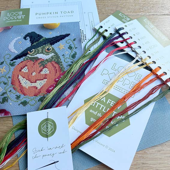 Cross stitch pattern with a pumpkin and witch hat, surrounded by embroidery threads and pattern sheets.