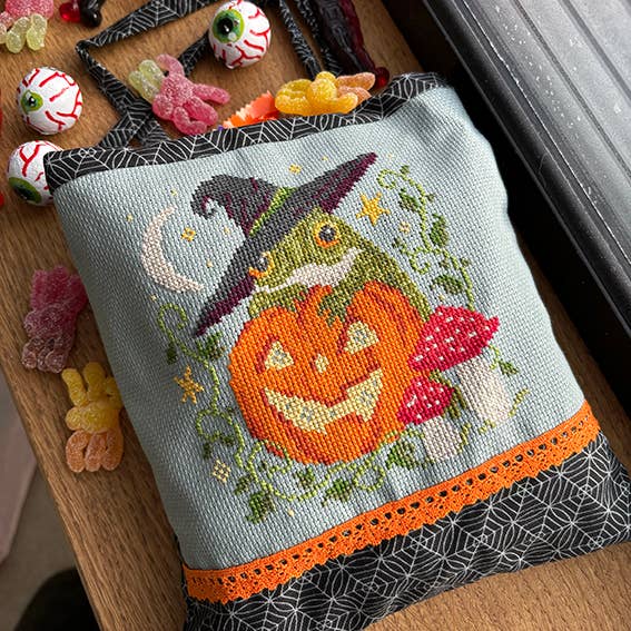 Halloween-themed cross-stitched pillow with a pumpkin and witch design on a wooden surface with candy.