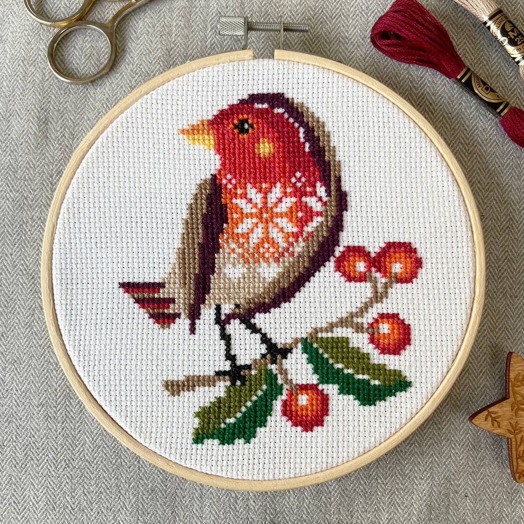 Cross-stitched robin on a branch with berries in an embroidery hoop, surrounded by cross-stitching supplies.