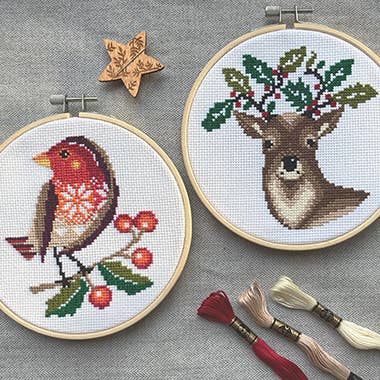 Two cross-stitched pieces on hoops with a robin and deer design, surrounded by embroidery tools on a gray background.