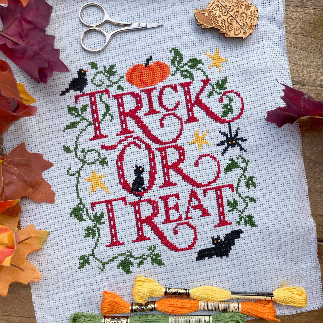 Cross-stitched Halloween design with 'Trick or Treat' text on fabric, surrounded by autumn leaves and embroidery supplies.