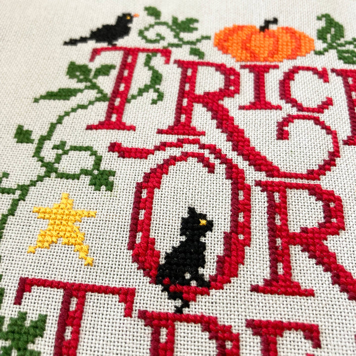 Cross-stitched design with 'Trick or Treat' text, black cat, pumpkin, and star on a white background.