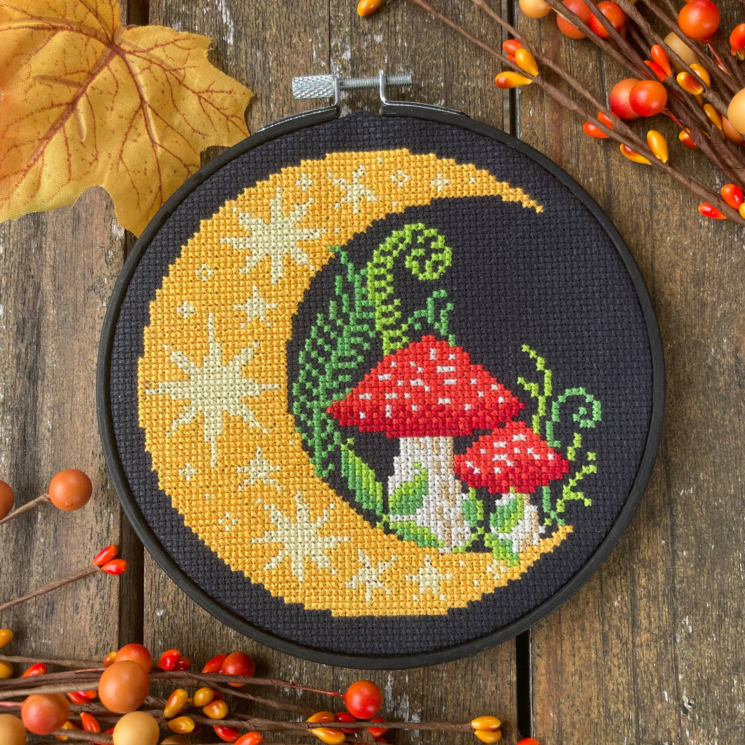 Cross-stitched design of mushrooms on a crescent moon with autumn leaves and berries in the background.