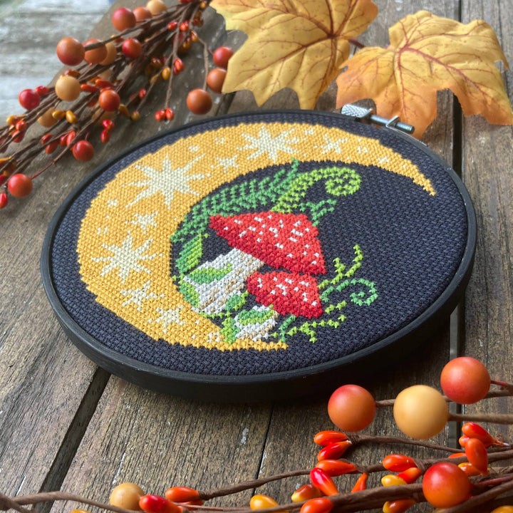 Cross-stitched design of a moon with red fabric and green leaves on a wooden surface with autumn leaves and berries.