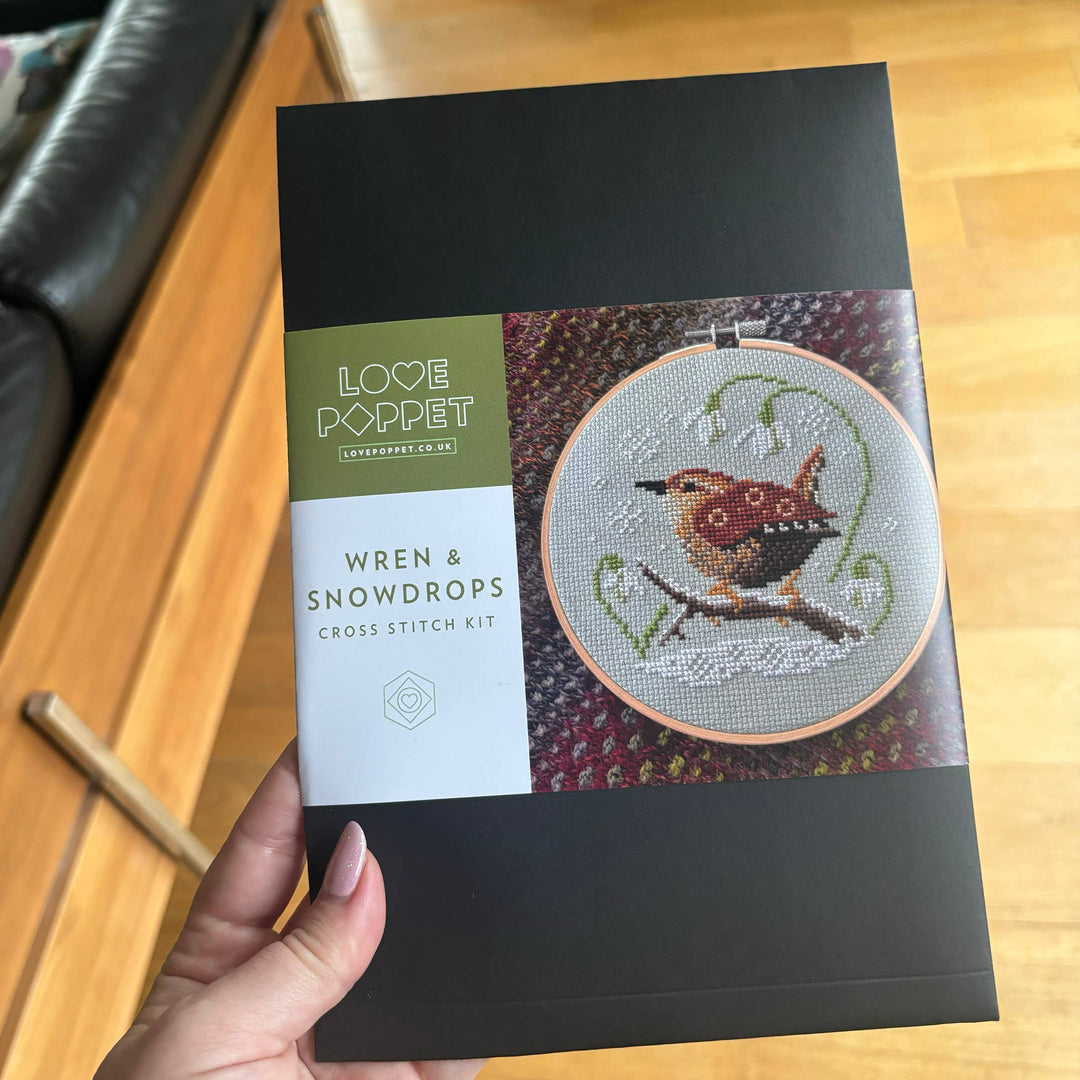 Cross stitch kit packaging with a wren and snowdrops design held by a hand on a wooden surface.