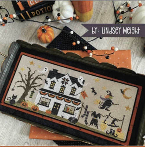Halloween-themed cross-stitch design with a haunted house and witch on a broomstick, displayed on a wooden surface.