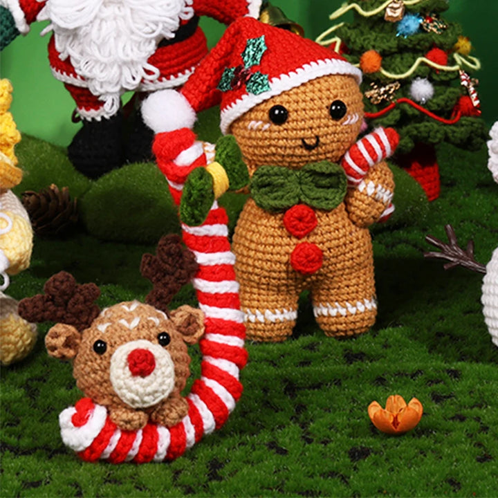 Crocheted gingerbread man and reindeer with candy cane on a green grass-like surface with Christmas tree in the background.