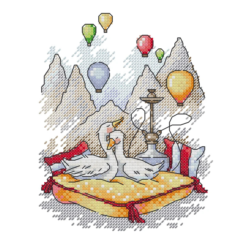 Cross-stitched design of two swans on a pillow with hot air balloons and mountains in the background.