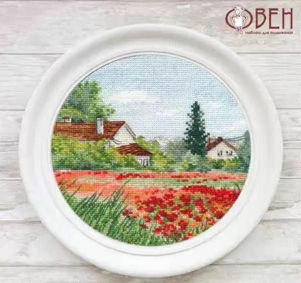Cross-stitched landscape with houses and a field of flowers in a white frame.