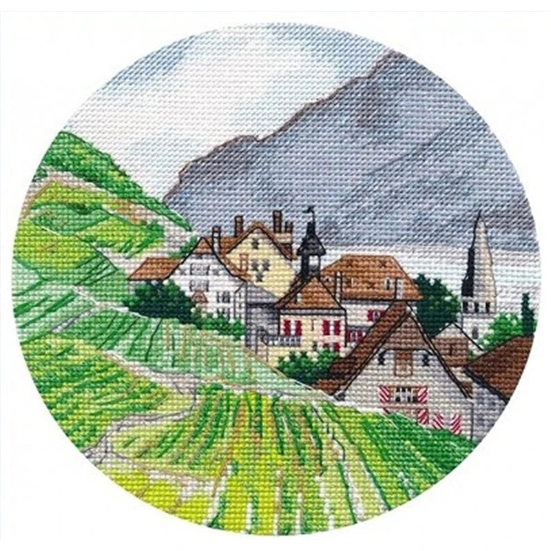 Circular needlepoint or cross-stitch design of a village with green fields and mountains.