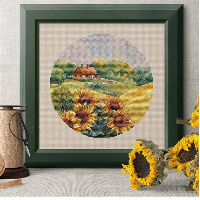Framed cross-stitched landscape with sunflowers and a house, surrounded by dried sunflowers.