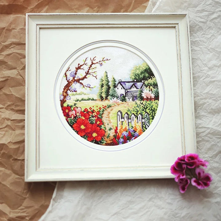 Framed cross-stitched landscape with flowers and house on a textured surface