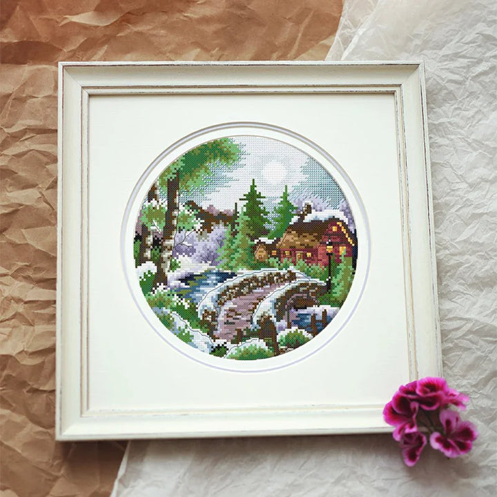 Framed cross-stitched landscape with a house and bridge on a textured surface.