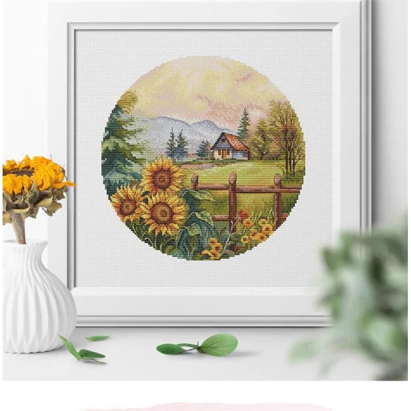 Framed cross-stitched landscape with sunflowers and a house on a white surface.