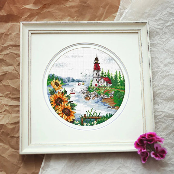 Framed cross-stitched picture of a lighthouse with sunflowers on a textured surface.