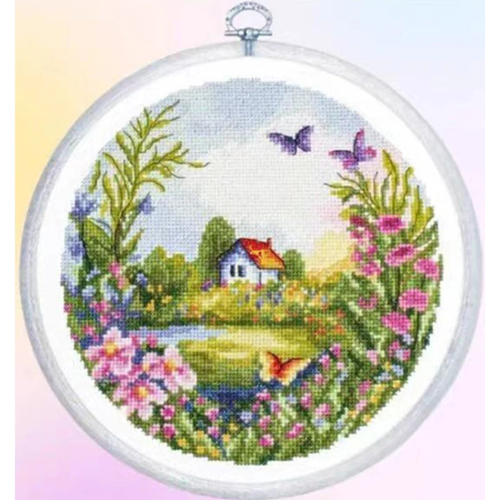 Cross-stitched landscape with house, flowers, and butterflies in an embroidery hoop.