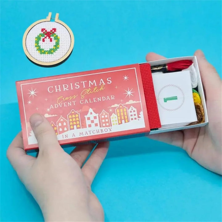 Christmas cross-stitch advent calendar in a matchbox held by hands on a blue background