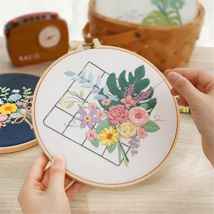 Embroidery hoop with floral design held by a person, with another hoop and materials in the background.