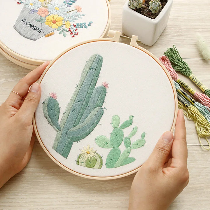 Embroidery hoop with cactus design held by hands on a wooden surface with embroidery threads and plants.