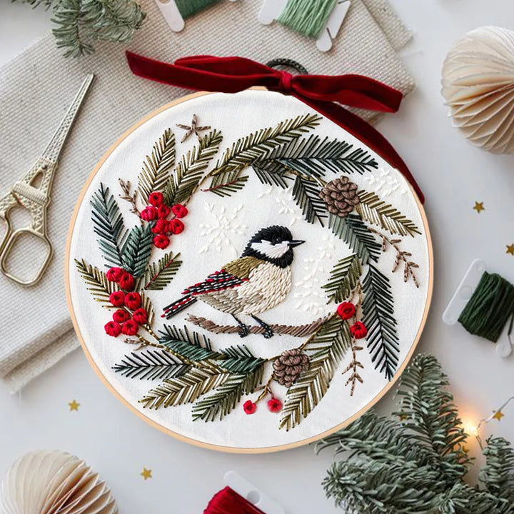 Embroidery hoop with a Christmas-themed design featuring a bird, berries, and foliage on a white surface.