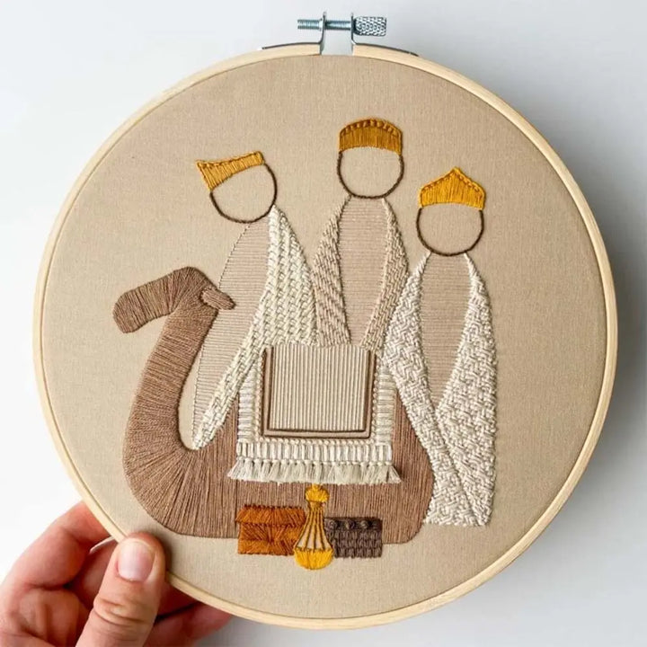 Embroidery hoop with a detailed embroidered design of three figures on beige fabric.