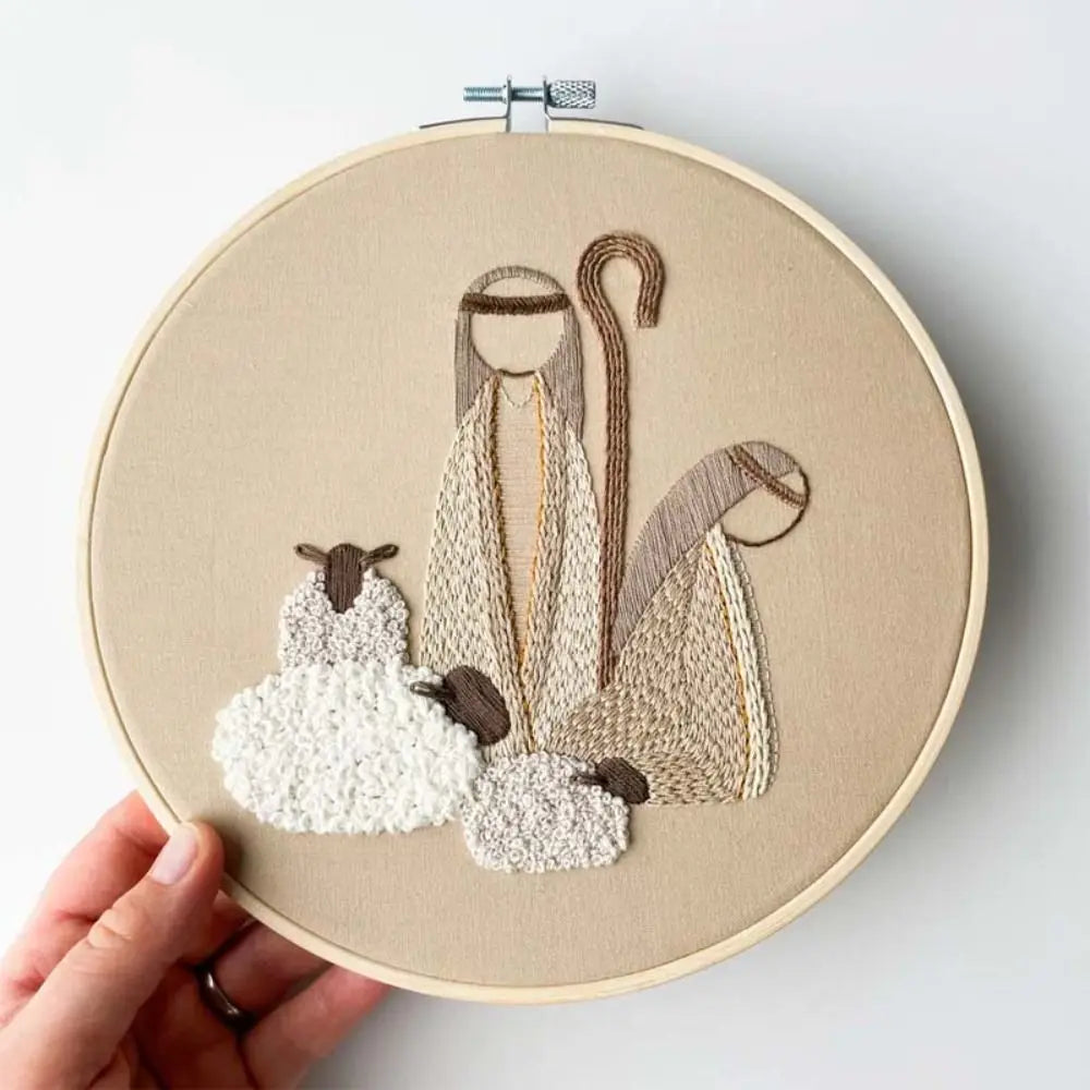 Embroidery hoop with a religious scene featuring a shepherd and two sheep on beige fabric.