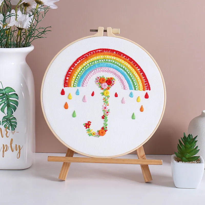 Embroidery hoop with a colorful rainbow and floral design on a stand against a beige wall.